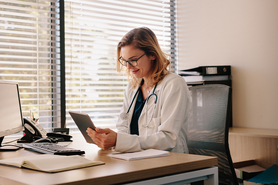 Employee Benefits - Female Doctor Working at Her Desk and Charting on Her Digital Tablet